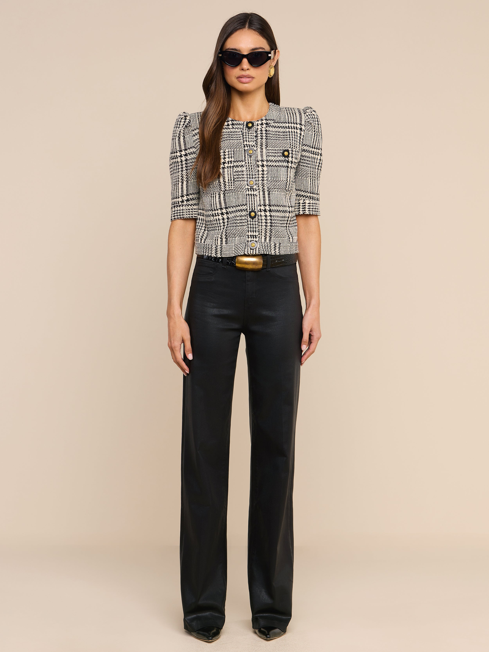 A woman wearing sunglasses and the Allegra Cropped Tweed Jacket layered over a short-sleeve plaid blouse, paired with black high-waisted pants and black shoes, stands against a plain white background, facing forward with a neutral expression.