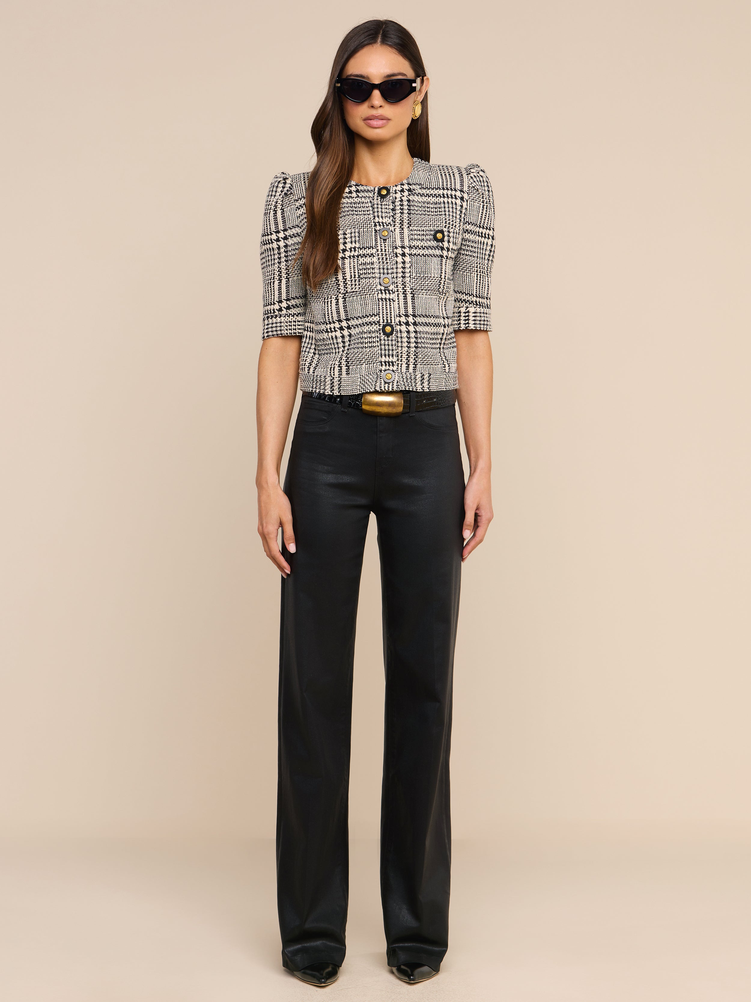 A woman wearing sunglasses and the Allegra Cropped Tweed Jacket layered over a short-sleeve plaid blouse, paired with black high-waisted pants and black shoes, stands against a plain white background, facing forward with a neutral expression.