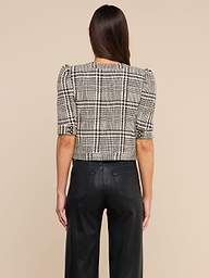 A woman stands facing away against a plain light background, wearing the Allegra Cropped Tweed Jacket—a black-and-white plaid tweed design with short sleeves—paired with black pants.