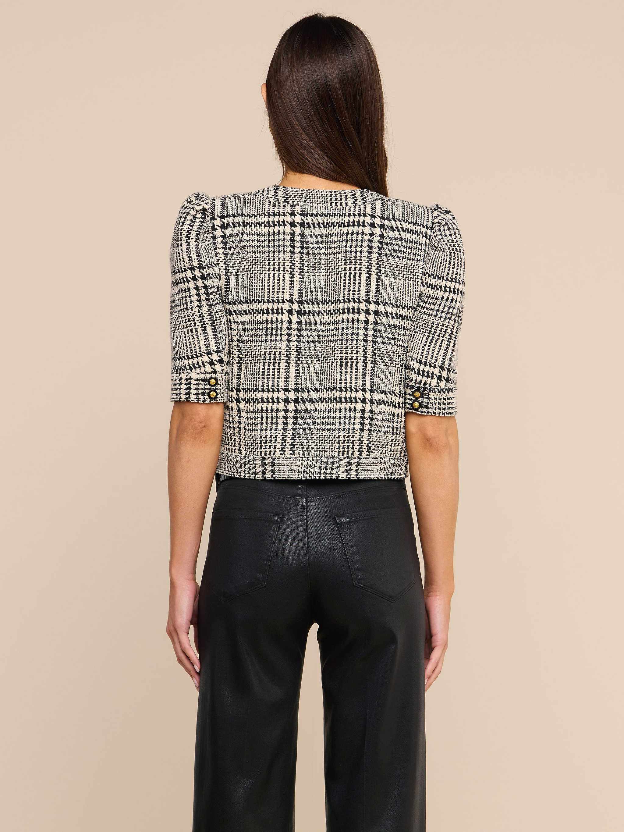 A woman stands facing away against a plain light background, wearing the Allegra Cropped Tweed Jacket—a black-and-white plaid tweed design with short sleeves—paired with black pants.