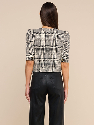 A woman, seen from behind, is wearing the Allegra Cropped Tweed Jacket—a black and white plaid tweed piece featuring gold buttons on the sleeves—paired with high-waisted black pants, set against a beige background.