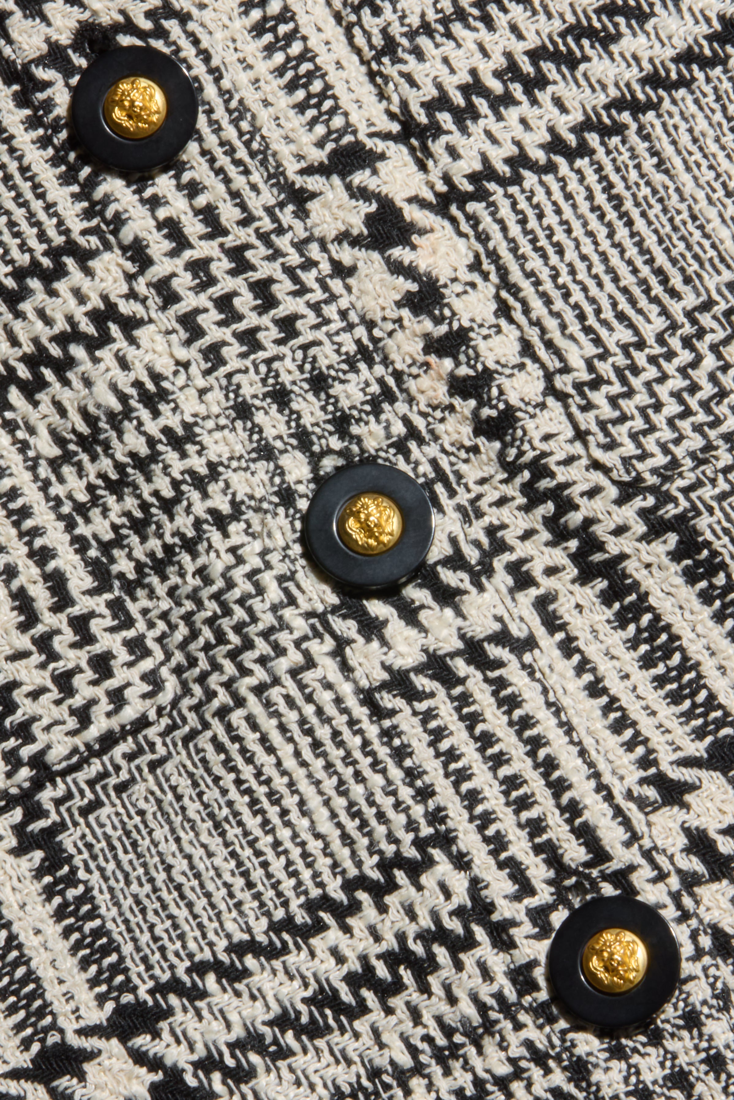 Close-up of black and white patterned fabric from the Allegra Cropped Tweed Jacket, featuring a round black button with a textured gold center. The fabric showcases zigzag and grid designs, creating a woven, textured appearance.