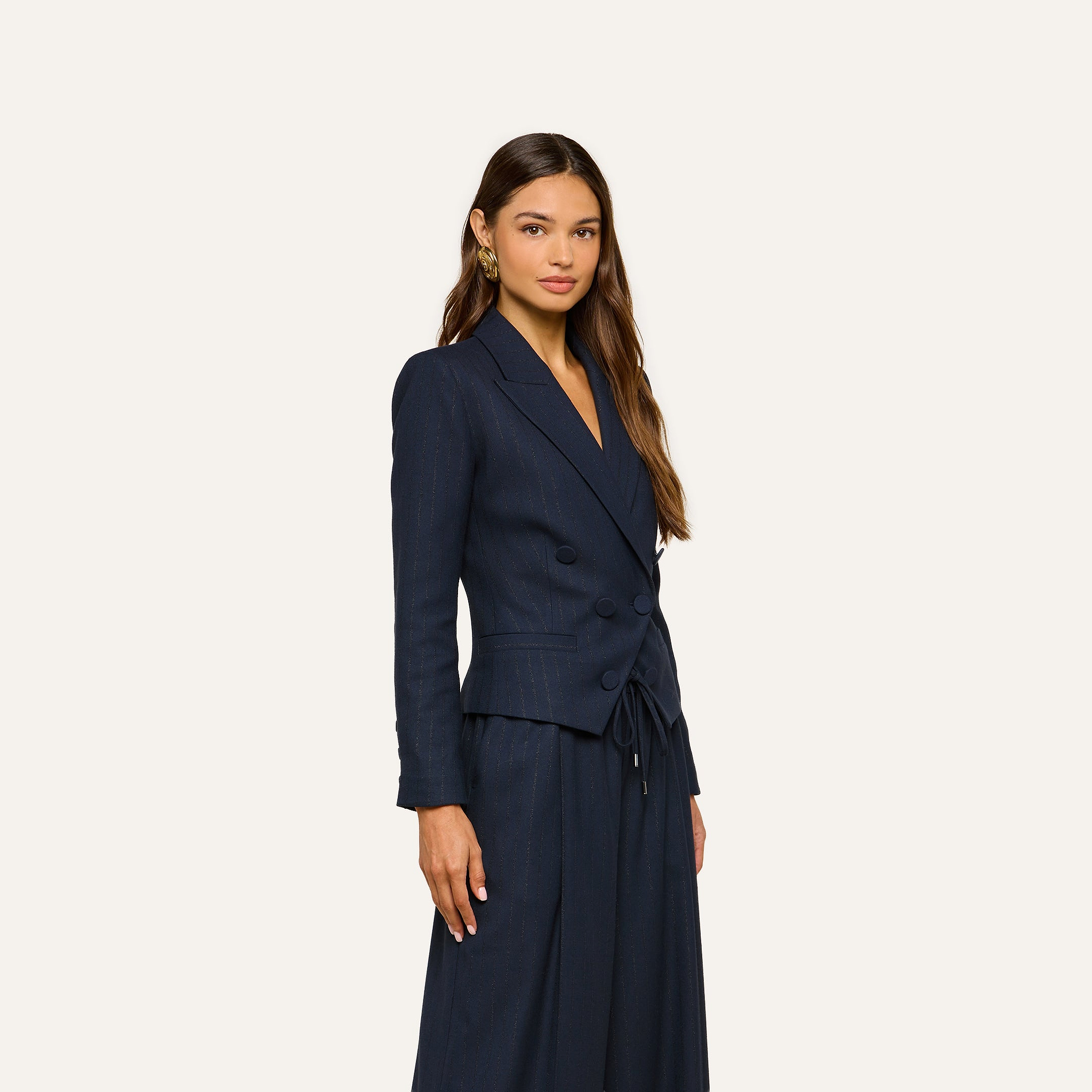 A woman poses against a plain light background, looking forward with a neutral expression, wearing the Risa Double-Breasted Blazer in navy blue with matching wide-leg pinstriped pants.