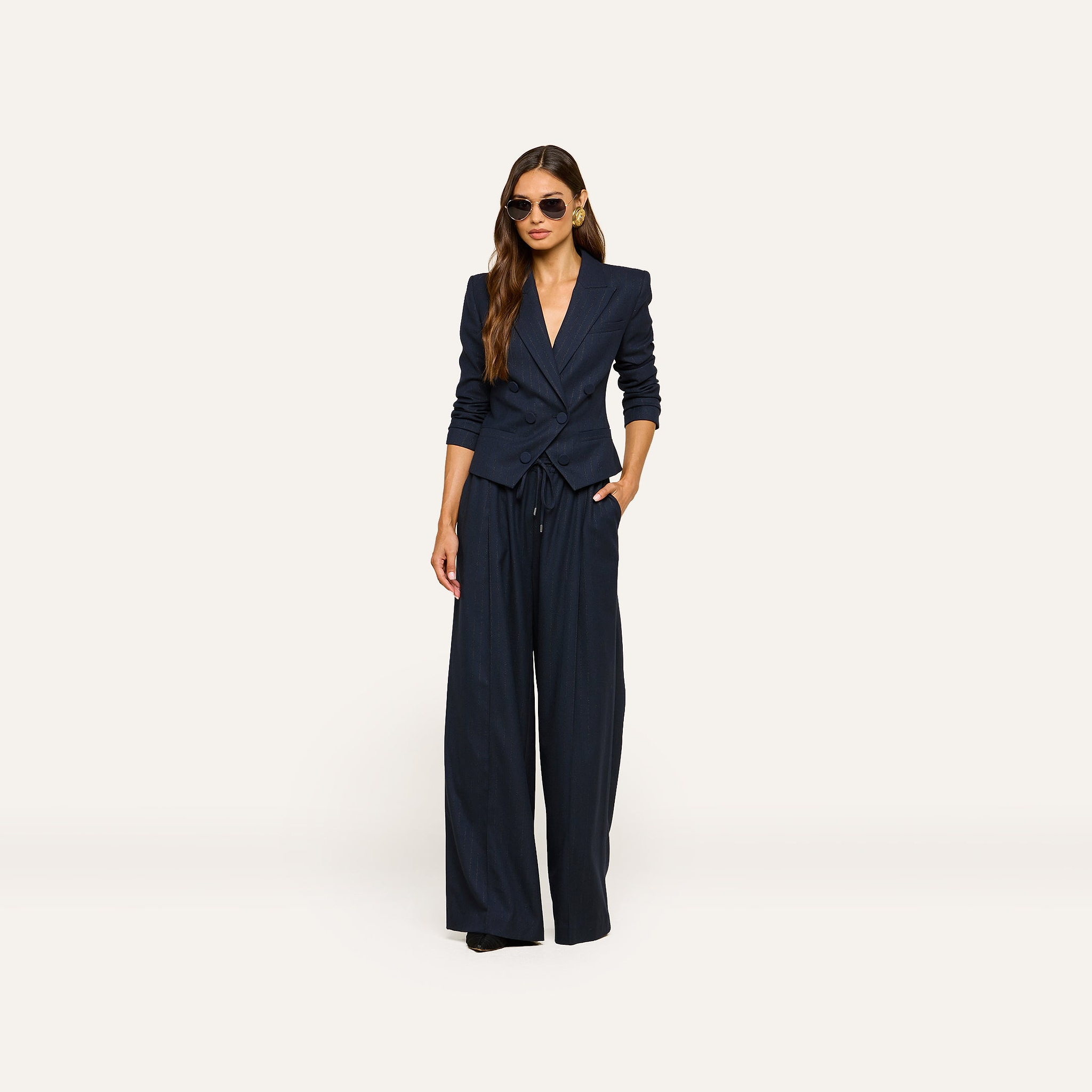 Model dressed in the Risa Double-Breasted Blazer in navy blue and matching wide-leg pants, one hand casually in her pocket, wearing sunglasses, and posing against a plain light background.