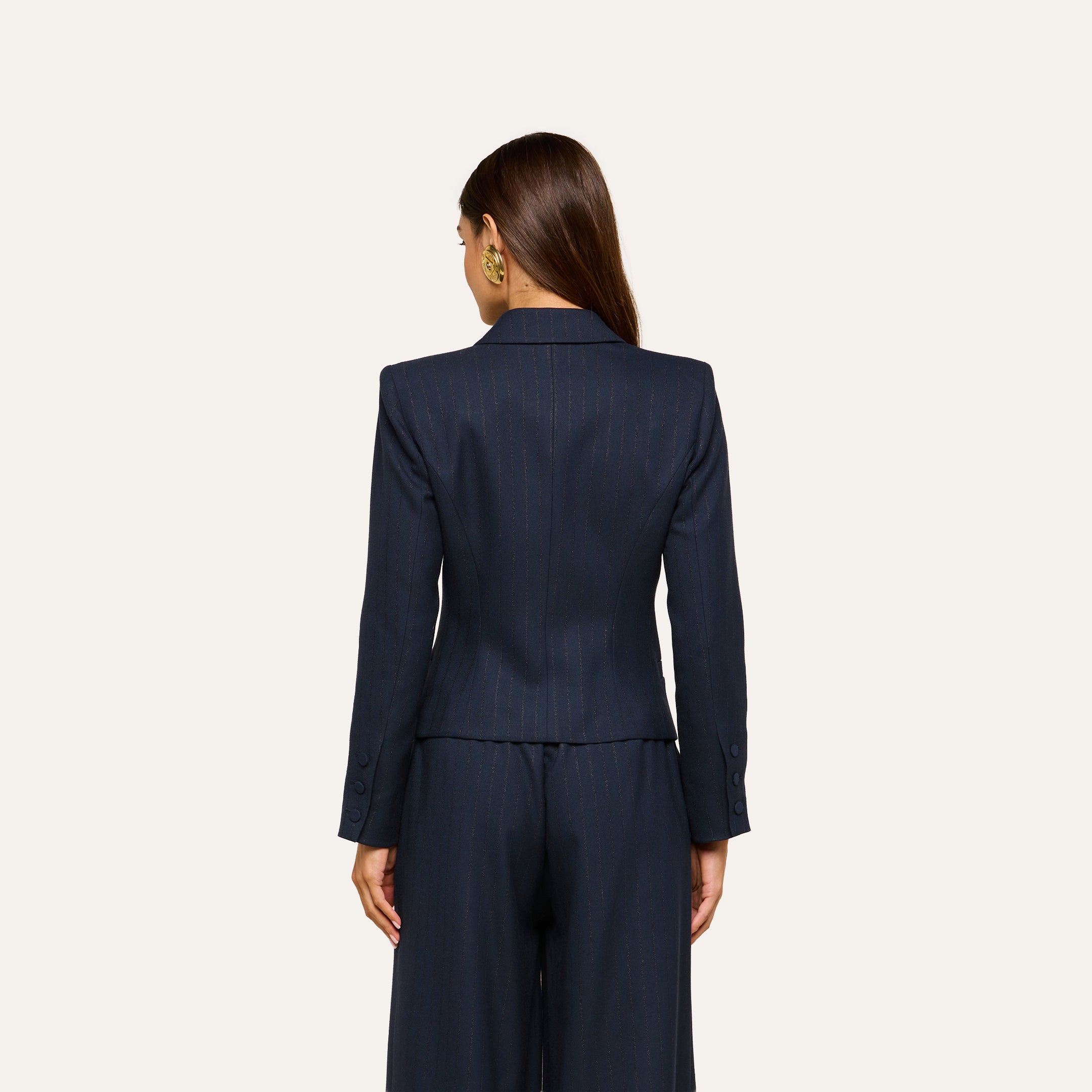 A woman with long brown hair stands facing away, wearing the Risa Double-Breasted Blazer and matching trousers against a plain light background.