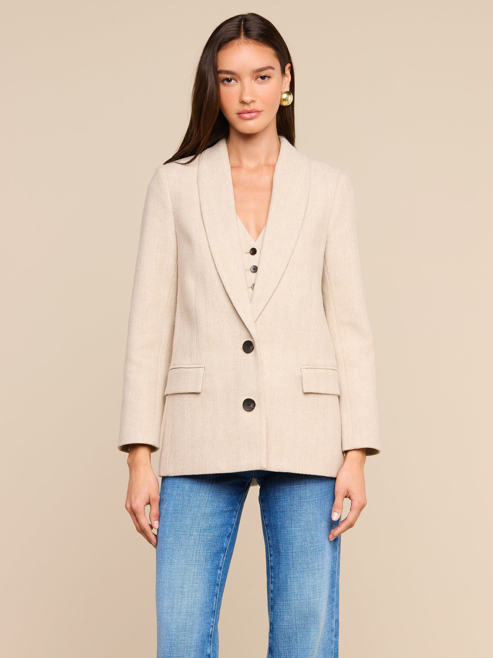 Channeling New York Fashion Week style, this woman wears the Amelie Blazer—a fitted beige blazer with black buttons—over a matching top, paired with blue jeans as she stands against a plain, light-colored background.