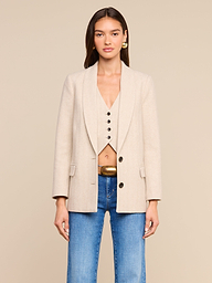 A woman stands facing forward wearing the Amelie Blazer, a fitted beige jacket paired with a matching buttoned vest and blue jeans. She has long brown hair, gold earrings, and a neutral expression, posed against a plain, light background.