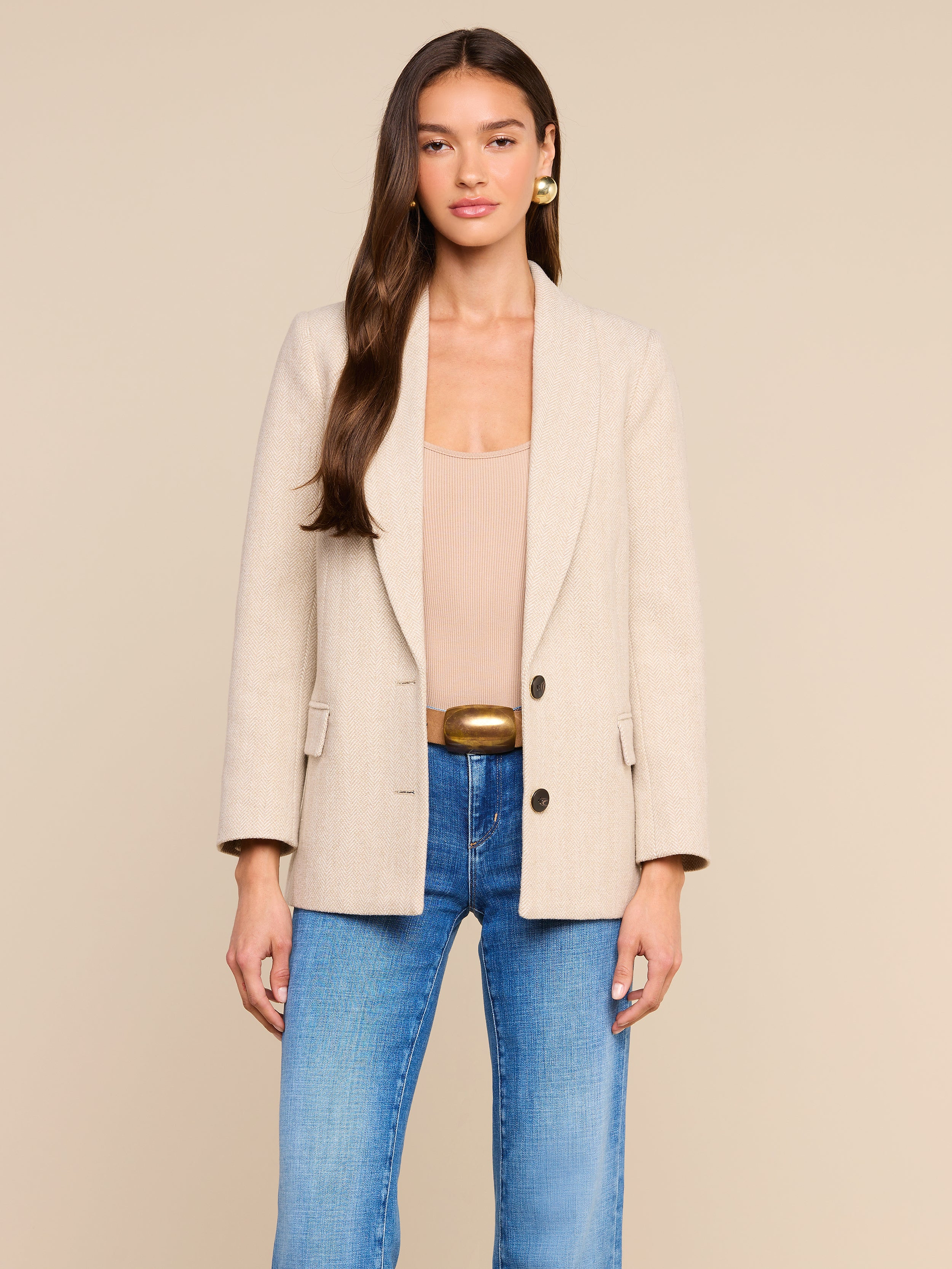 A woman with long brown hair wears the Amelie Blazer—a fitted beige blazer—over a nude top and blue jeans, standing against a plain light background. She faces forward with a neutral expression, her hands relaxed by her sides.