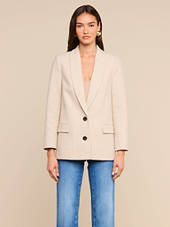 A woman stands facing forward, wearing the Amelie Blazer—a fitted beige herringbone tweed jacket with two black buttons and front pockets—paired with blue jeans and gold earrings, against a plain light background.