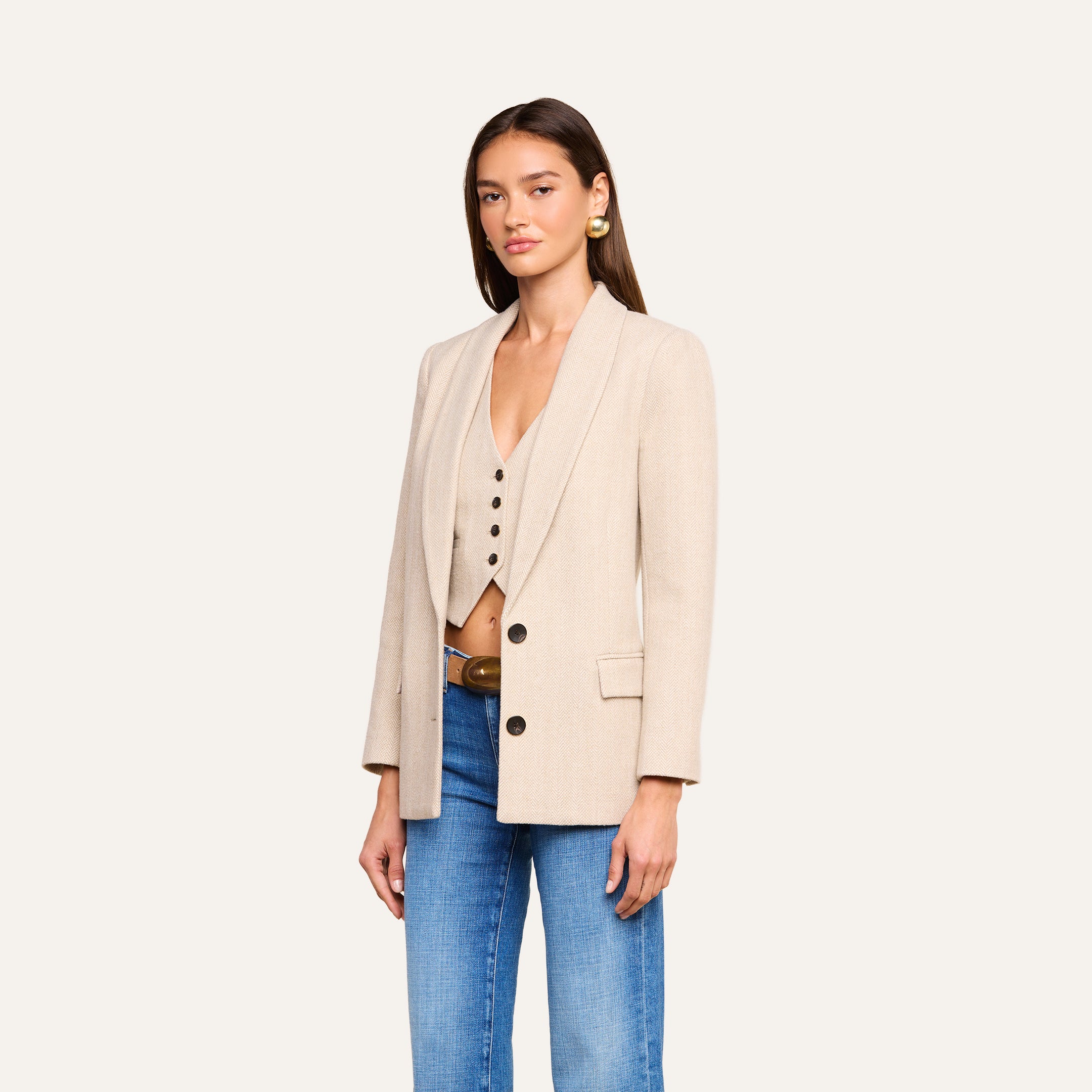 Sure Here’s a revised version incorporating the product name: A woman wears the Amelie Blazer—a beige fitted style—over a coordinating vest and blue jeans, standing against a plain background. With long straight hair, gold earrings, and a neutral expression, she exudes an effortless look reminiscent of New York Fashion Week.