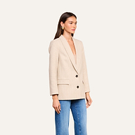A woman wearing the Amelie Blazer—a fitted beige jacket with two black buttons—over a matching top and blue jeans stands against a plain, light background, evoking the polished style seen at New York Fashion Week.