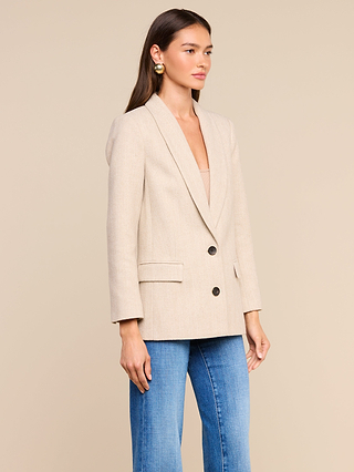 Wearing the Amelie Blazer—a fitted beige jacket featuring two buttons and flap pockets—a woman with long brown hair pairs it with blue jeans and gold earrings, standing against a plain beige background in a look inspired by the refined styles of New York Fashion Week.
