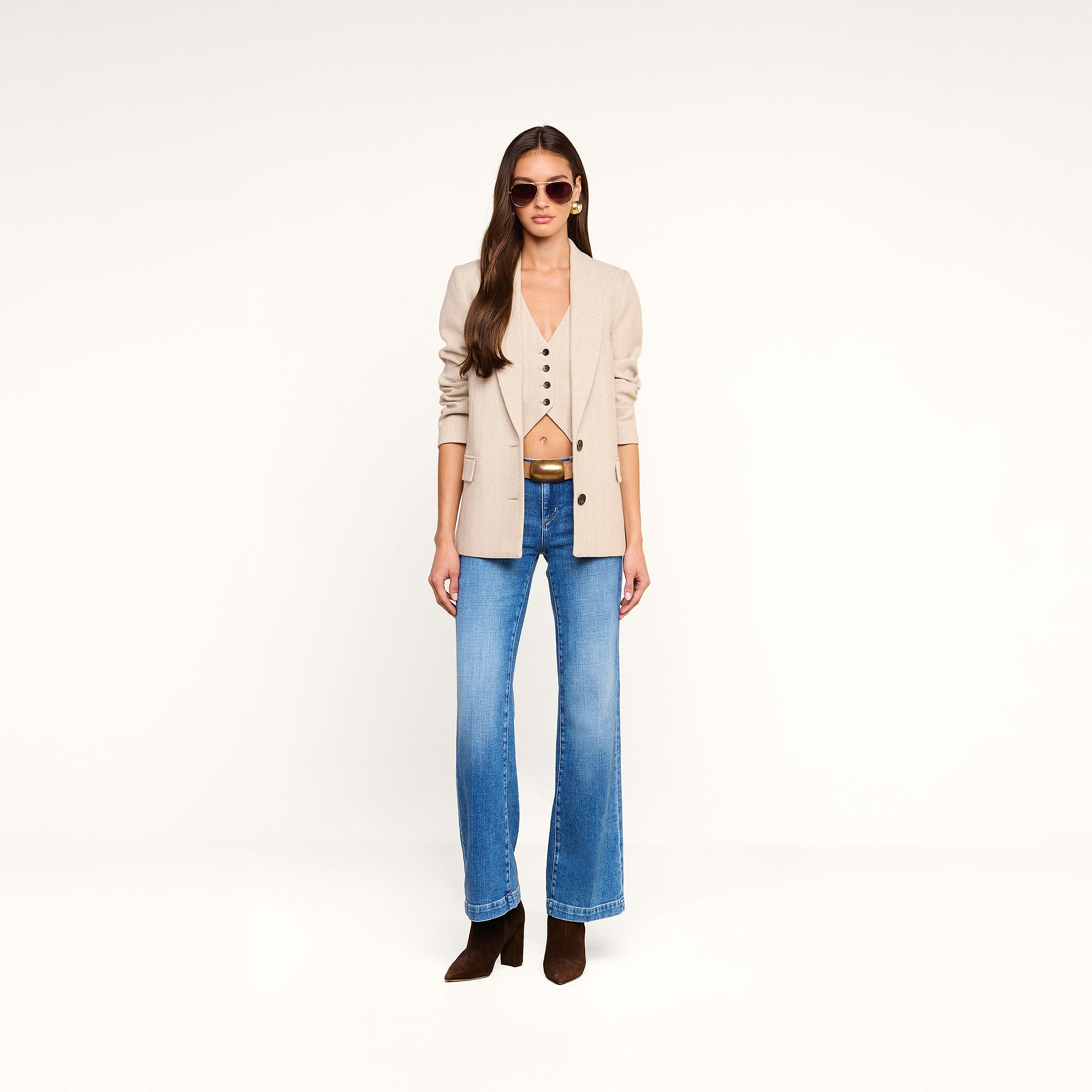 A woman with long hair wearing sunglasses, the Amelie Blazer in beige over a matching vest, blue jeans, and brown boots stands against a plain white background.