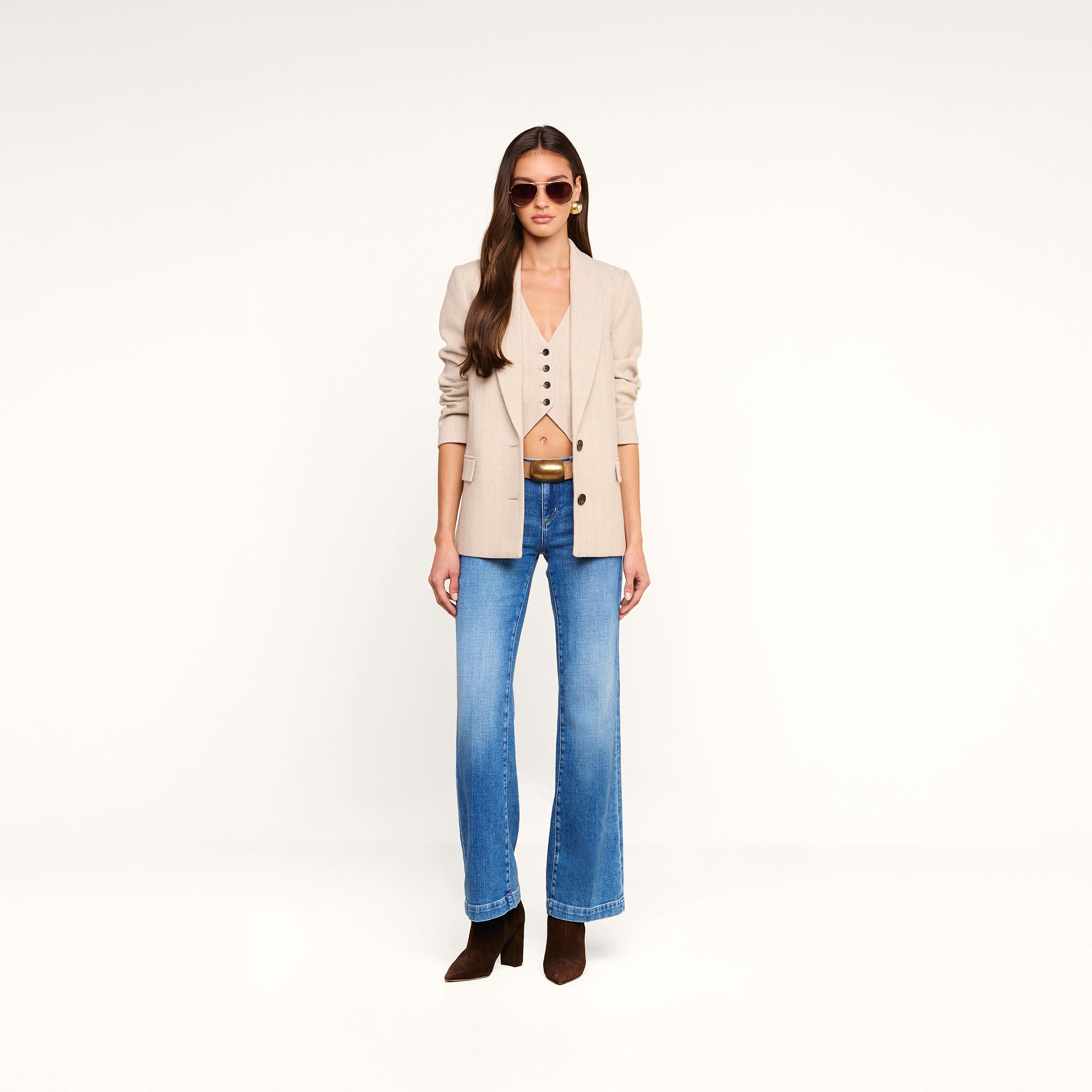 A woman with long hair wearing sunglasses, the Amelie Blazer in beige over a matching vest, blue jeans, and brown boots stands against a plain white background.