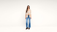 Woman wearing sunglasses, a beige blazer, a tan top, blue wide-leg jeans, and brown boots standing against a plain white background.