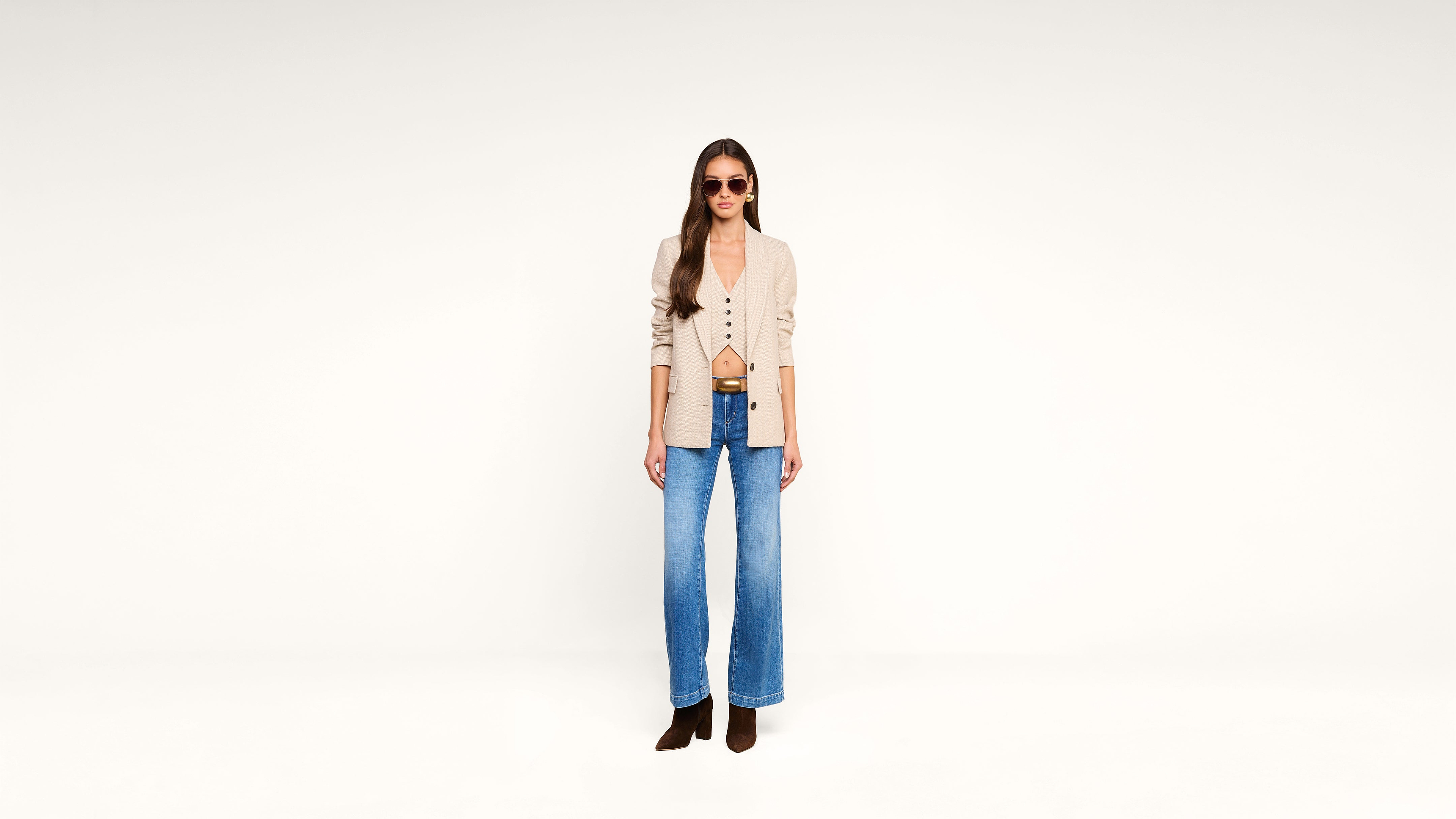 Woman wearing sunglasses, a beige blazer, a tan top, blue wide-leg jeans, and brown boots standing against a plain white background.
