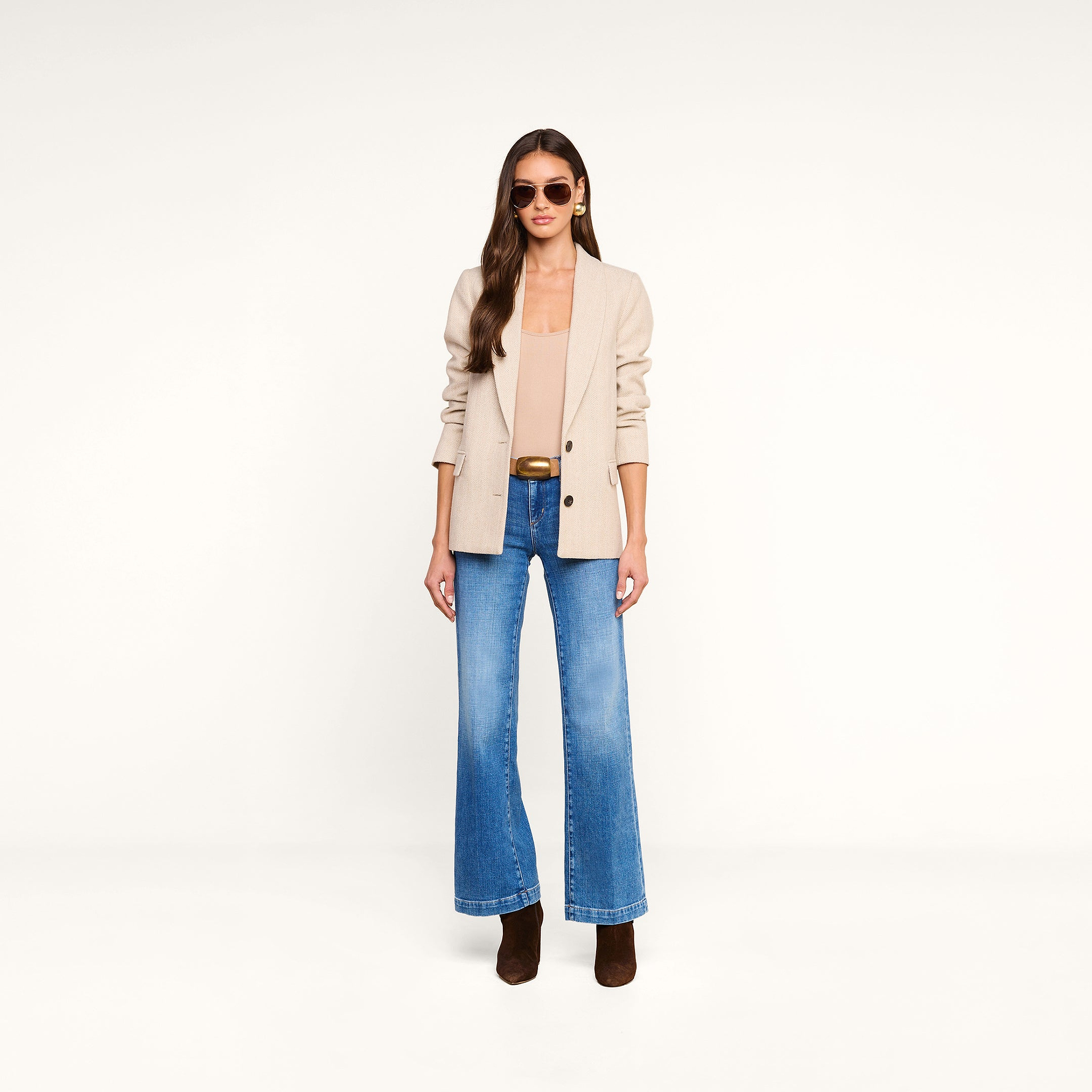 A woman wearing the Amelie Blazer, a tan top, blue wide-leg jeans, brown boots, sunglasses, and gold earrings stands against a plain white background.