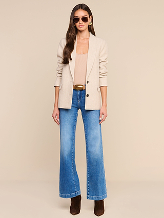 Una donna è in piedi su uno sfondo beige a tinta unita, con lo sguardo rivolto dritto davanti a sé e le mani lungo i fianchi; indossa il blazer Amelie beige sopra un top color cuoio, jeans a zampa blu, una cintura marrone, stivali marroni e occhiali da sole: un look che ricorda lo stile della Settimana della Moda di New York.