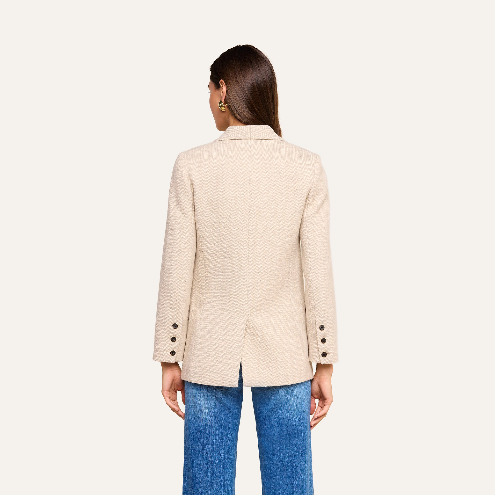 A woman with straight brown hair is shown from the back wearing the fitted Amelie Blazer in beige, featuring button details on the sleeves, paired with blue jeans against a plain light background.