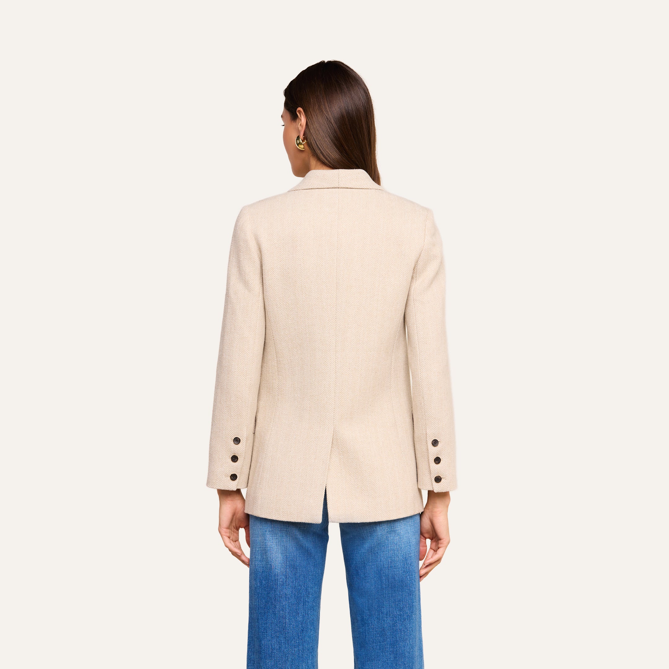 A woman with straight brown hair is shown from the back wearing the fitted Amelie Blazer in beige, featuring button details on the sleeves, paired with blue jeans against a plain light background.