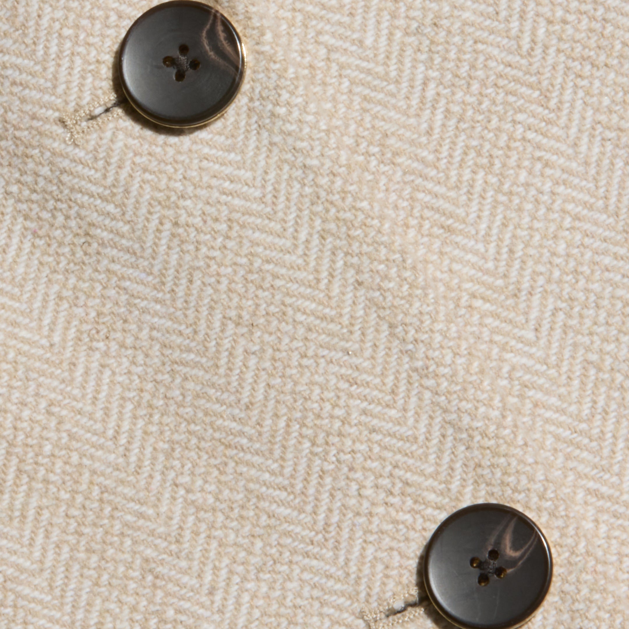 Close-up of cream-colored herringbone tweed fabric with two dark, round, four-hole buttons sewn onto it. The texture and weave of the fabric are clearly visible, embodying the tailored elegance of the Amelie Blazer—a fitted piece inspired by New York Fashion Week.