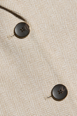 Nahaufnahme eines beigen Tweedstoffs im Fischgrätenmuster mit zwei parallel angenähten dunkelbraunen Knöpfen. Die Struktur und die Nähdetails des Amelie-Blazers sind deutlich zu erkennen.