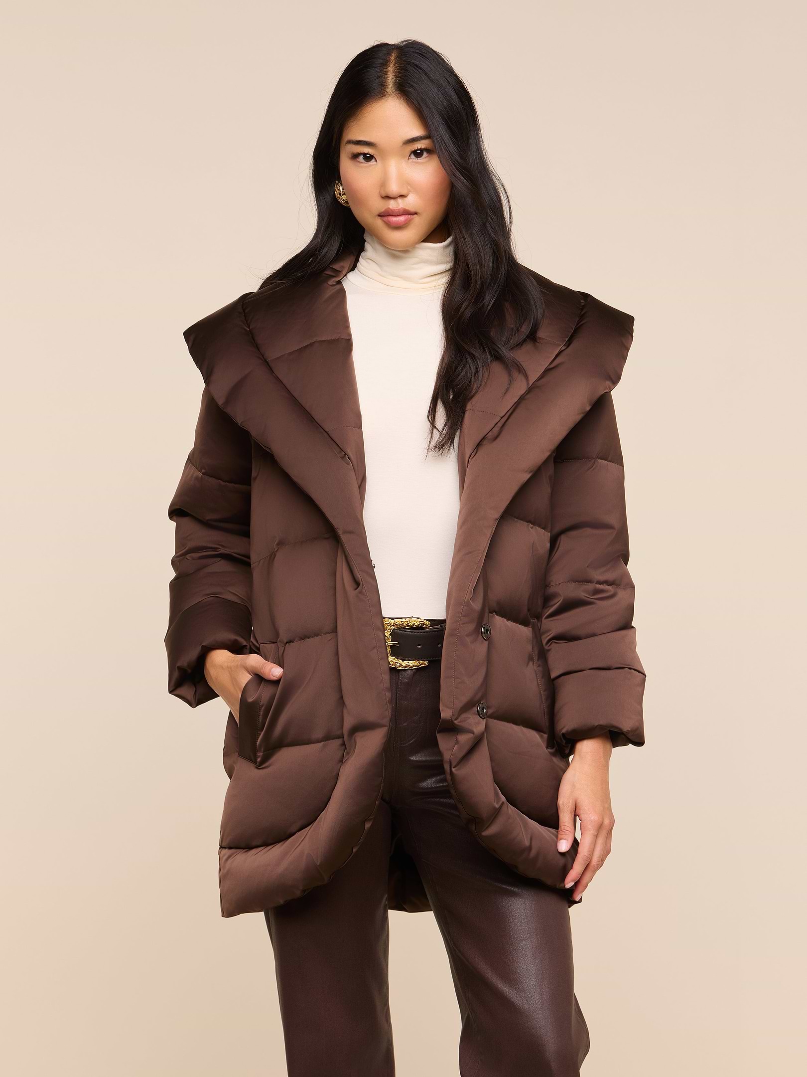 A woman with long dark hair wears the Glacier Shawl Puffer Jacket in brown, layered over a cream turtleneck and paired with brown pants. She stands with one hand in her jacket pocket against a plain light background.