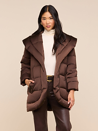 A woman with long dark hair wears the Glacier Shawl Puffer Jacket in brown, layered over a cream turtleneck and paired with brown pants. She stands with one hand in her jacket pocket against a plain light background.