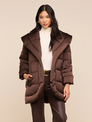 A woman stands facing forward against a beige background, her hands tucked into the pockets of the Glacier Shawl Puffer Jacket—a water-repellent brown puffer—layered over a cream turtleneck and brown pants.