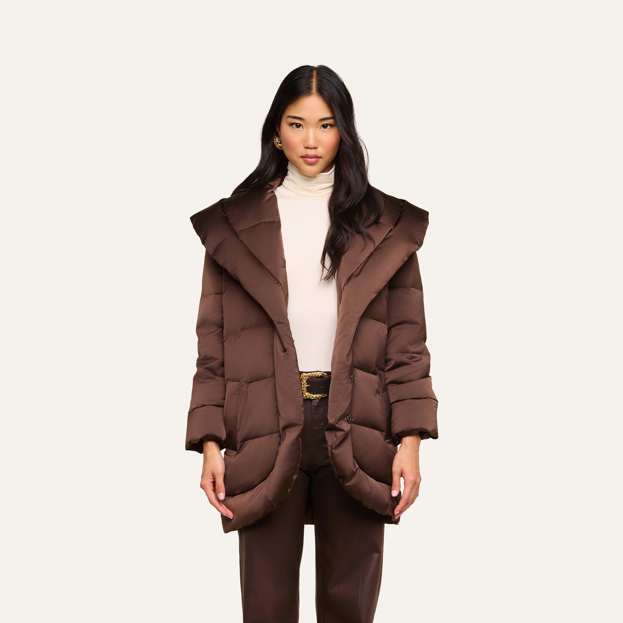 A woman wearing the Glacier Shawl Puffer Jacket—a brown water-repellent puffer with a wide collar—paired with a cream turtleneck and brown pants, stands against a plain light background.
