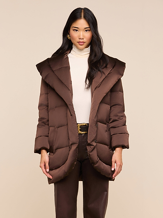 A woman stands facing forward against a plain beige background, wearing the Glacier Shawl Puffer Jacket in brown with a large collar, paired with a cream turtleneck and brown pants.