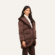 A woman models the Glacier Shawl Puffer Jacket—a brown, hooded puffer—paired with a white turtleneck and brown pants. She stands against a plain light background, facing the camera with a neutral expression.