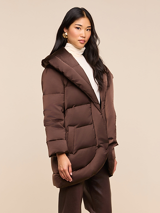 A woman with long dark hair wears the Glacier Shawl Puffer Jacket, featuring a water-repellent brown design, paired with a cream turtleneck and statement earrings. She stands against a plain beige background, looking confidently at the camera.