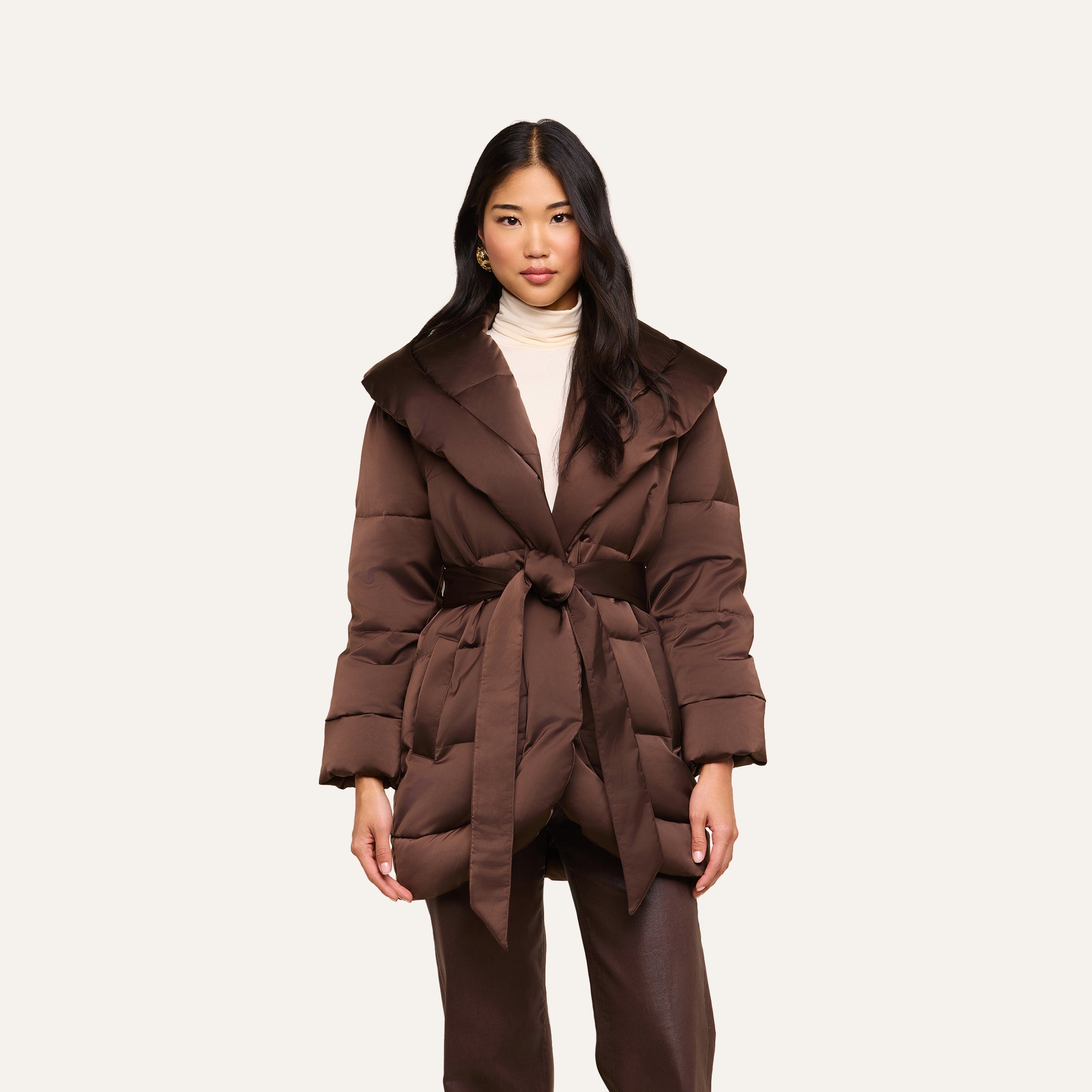 A woman stands facing forward against a plain light background, wearing the Glacier Shawl Puffer Jacket—a brown, belted, water-repellent jacket—layered over a light turtleneck and paired with brown pants.