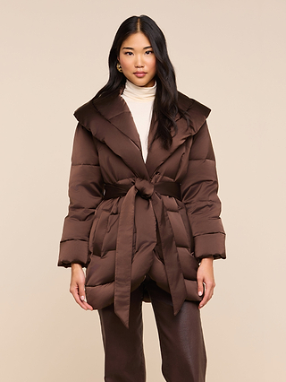 A woman wearing the Glacier Shawl Puffer Jacket, belted at the waist over a cream turtleneck and brown pants, stands against a beige background.