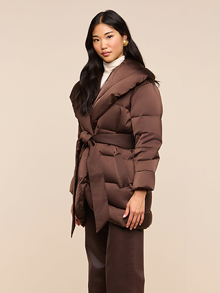 A woman with long black hair wears the Glacier Shawl Puffer Jacket, featuring a brown belted design, layered over a white turtleneck and brown pants while standing against a plain beige background. The jacket’s water-repellent finish brings both style and practical appeal to her look.