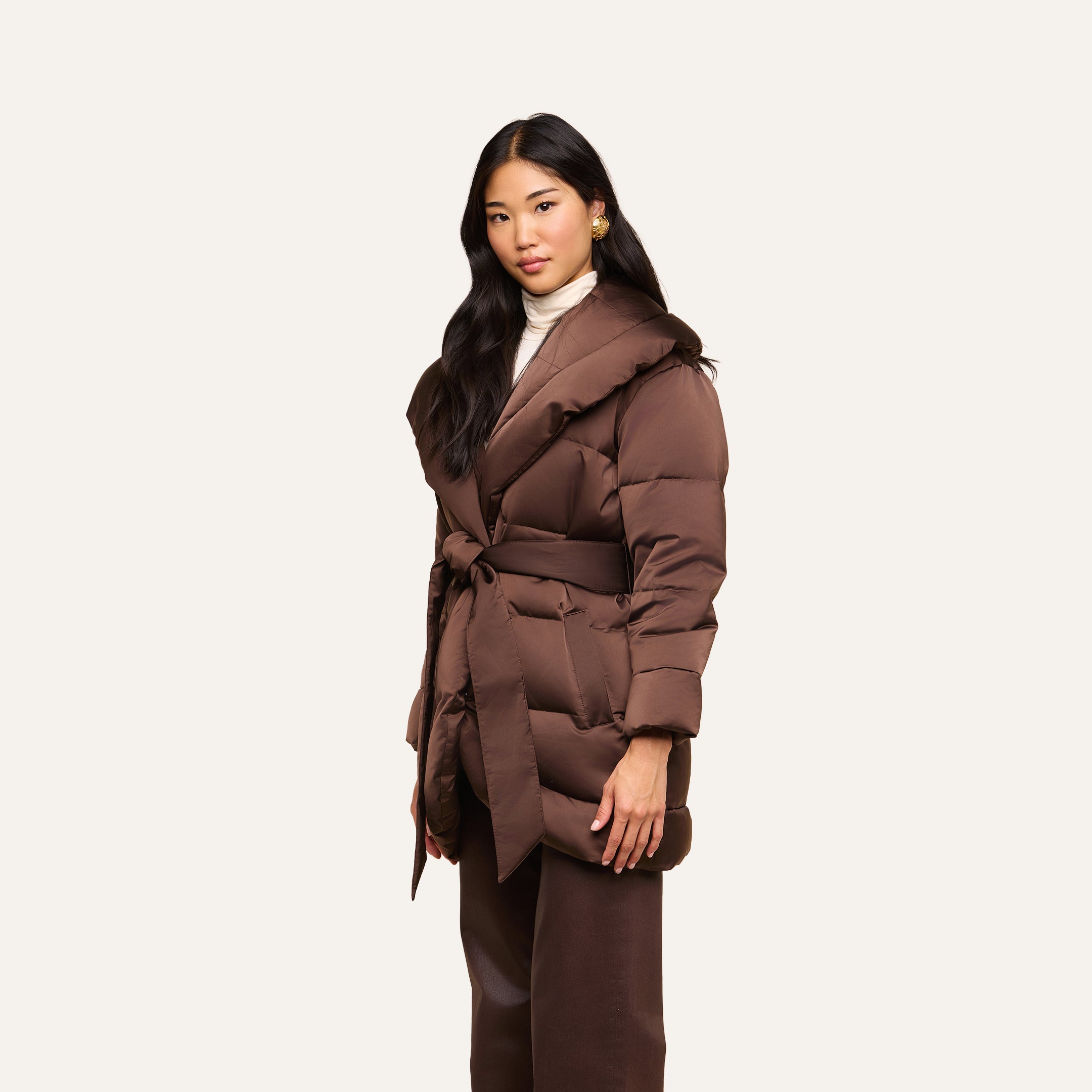 A woman with long dark hair wears the Glacier Shawl Puffer Jacket, styled over a cream turtleneck and brown pants, standing against a plain light background.