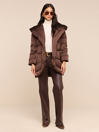 A woman with long dark hair wears the Glacier Shawl Puffer Jacket in brown over a cream turtleneck, paired with brown pants and shoes. She completes the look with large round sunglasses and poses against a plain beige background.