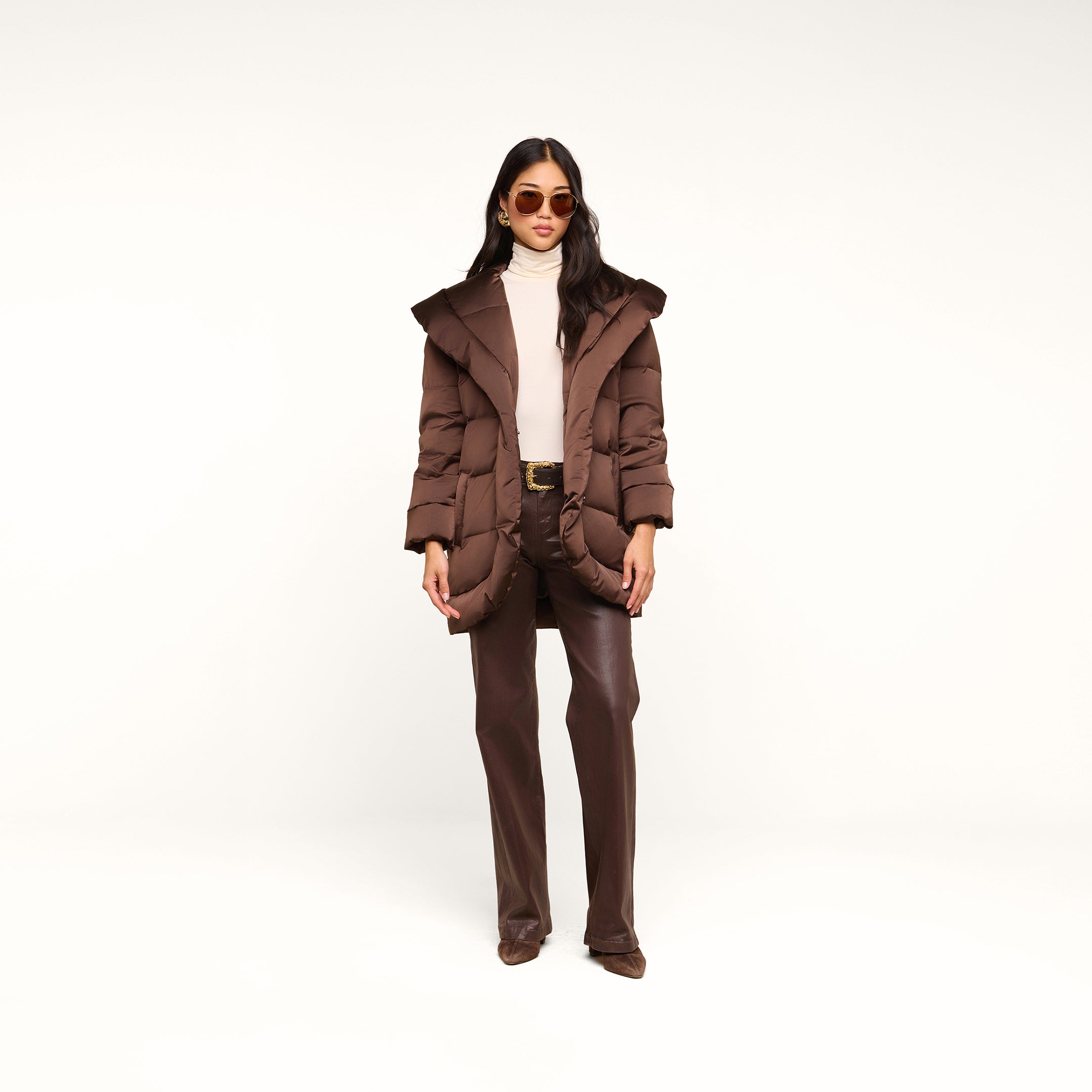 A woman wearing the Glacier Shawl Puffer Jacket in brown, paired with a white turtleneck, brown pants, and sunglasses, stands against a plain white background.