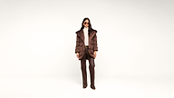 A woman stands against a plain white background, wearing sunglasses, a brown puffer coat, taupe turtleneck, brown pants, and brown shoes, with her hands in her pockets.