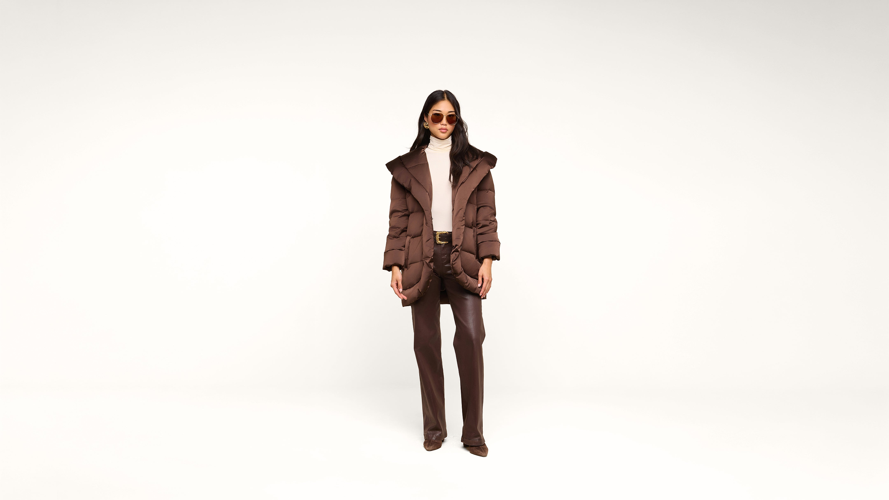 A woman stands against a plain white background, wearing sunglasses, a brown puffer coat, taupe turtleneck, brown pants, and brown shoes, with her hands in her pockets.