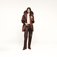 A woman stands against a plain white background wearing the Glacier Shawl Puffer Jacket, paired with brown pants, a beige top, and brown shoes, as she holds the jacket’s collar.