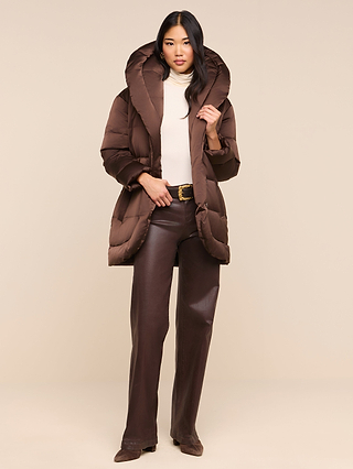 A woman stands against a neutral background wearing the Glacier Shawl Puffer Jacket—a brown, water-repellent puffer with a large hood—paired with an ivory turtleneck, brown wide-leg pants, and brown shoes.