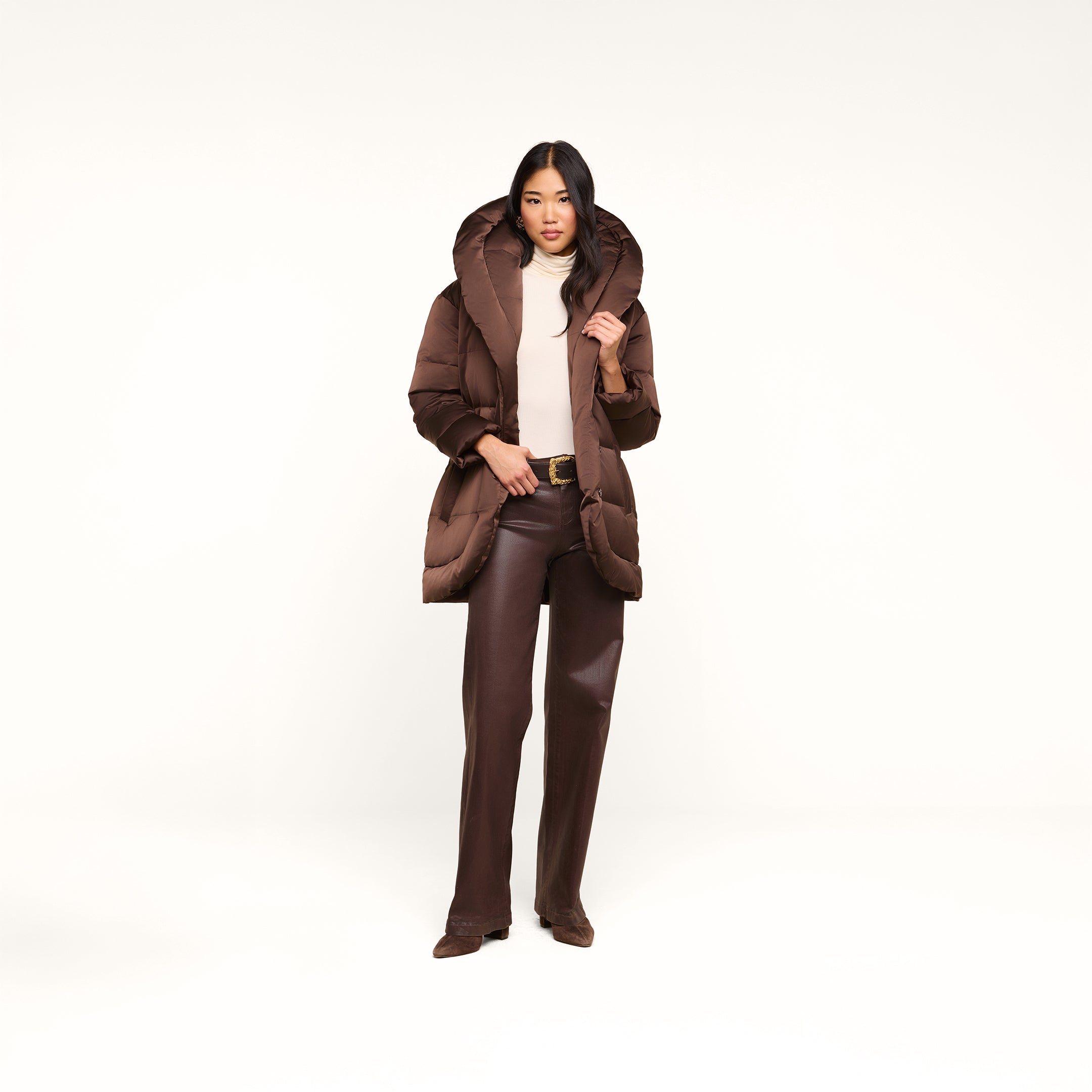 A woman stands against a plain white background wearing the Glacier Shawl Puffer Jacket, paired with brown pants, a beige top, and brown shoes, as she holds the jacket’s collar.