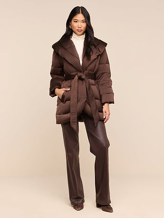 A woman stands against a plain light beige background wearing the Glacier Shawl Puffer Jacket—a brown, water-repellent belted coat—layered over a cream turtleneck and brown pants, paired with brown shoes.