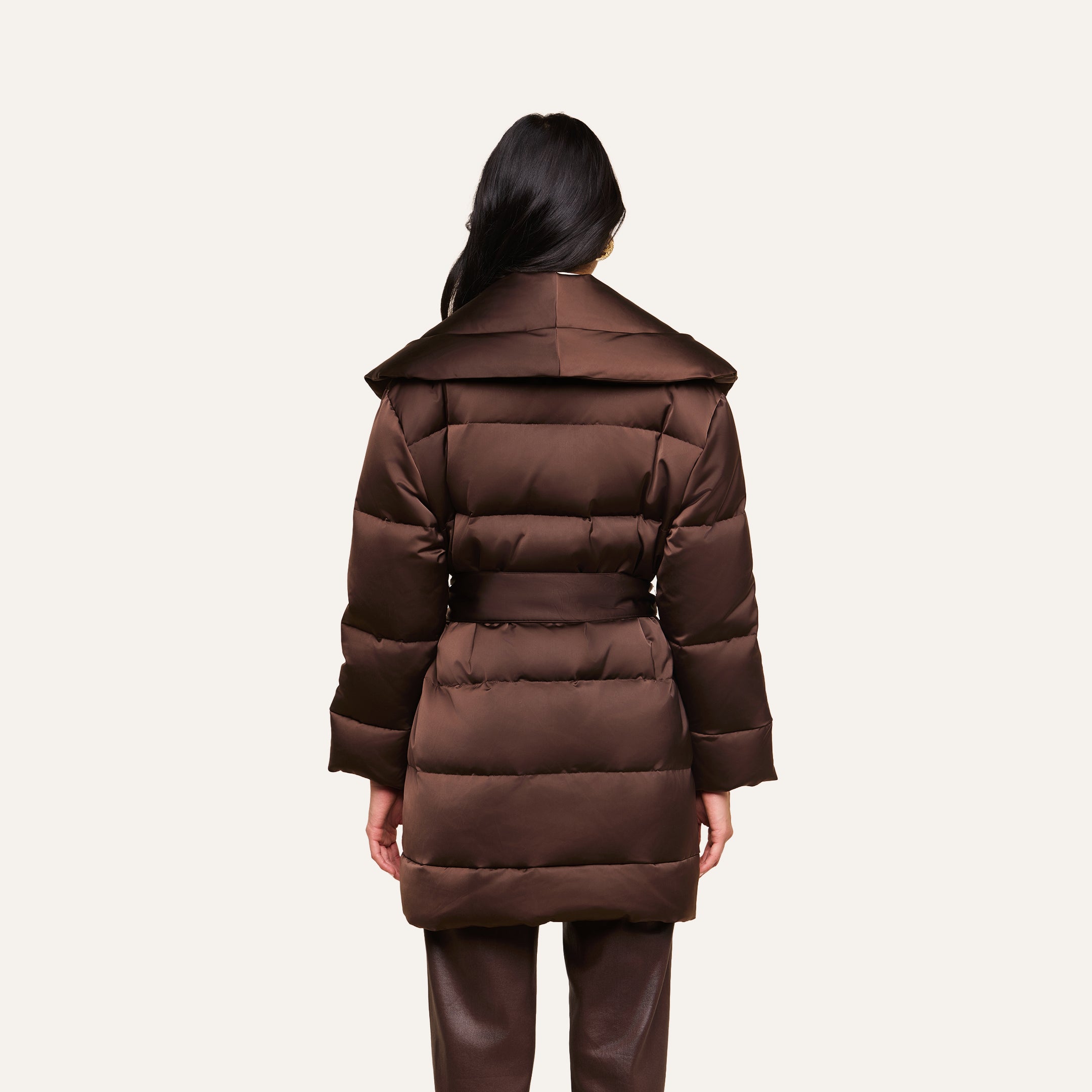 A person with long dark hair, seen from behind, is wearing the Glacier Shawl Puffer Jacket—a brown belted puffer jacket with a wide collar—paired with brown pants, and standing against a plain light background.