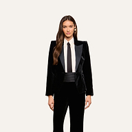 A woman wearing the Philippa Velvet Blazer with satin lapels, styled with a white dress shirt, black tie, and black cummerbund, stands against a plain light background. She has long, wavy hair and gazes forward.