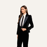 A woman with long brown hair stands facing left against a plain off-white background, wearing the Philippa Velvet Blazer with satin lapels, styled over a white button-up shirt, black tie, and matching black pants. She accessorizes her look with large round earrings.