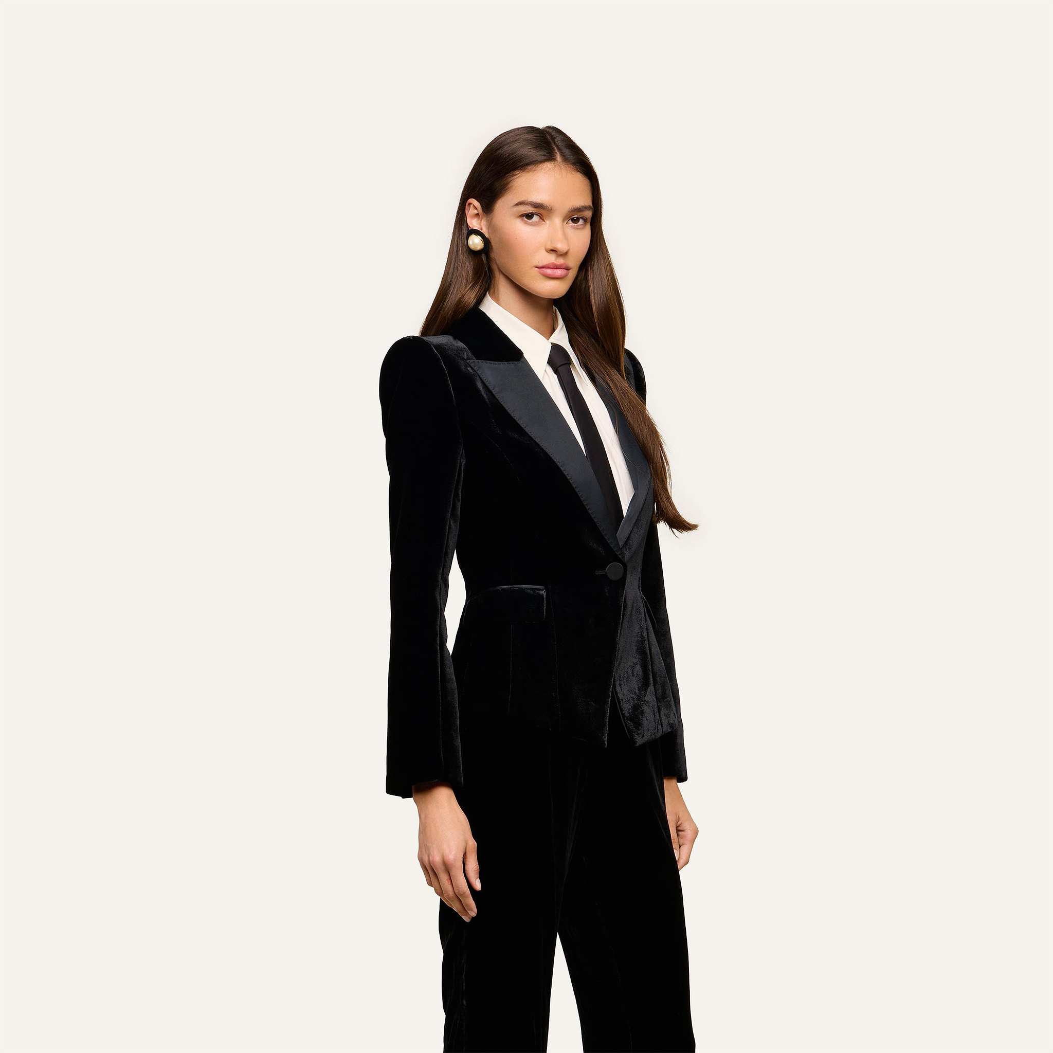 A woman wearing the Philippa Velvet Blazer, featuring black velvet fabric and satin lapels, along with a white shirt and a black tie, stands against a plain light background. She has long brown hair and is accessorized with large earrings.