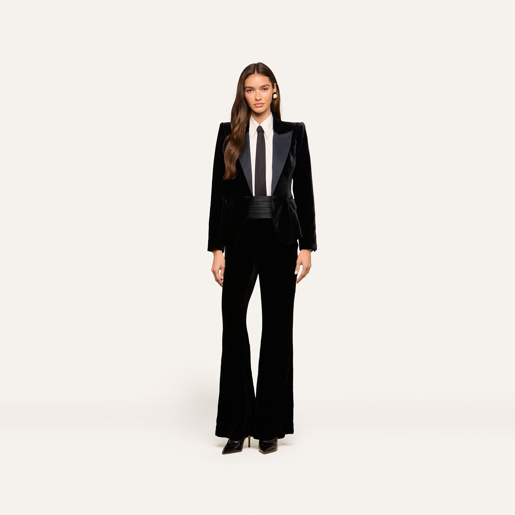 A woman stands facing forward, wearing the Philippa Velvet Blazer with satin lapels over a white shirt and black tie. She has long, loose hair and is wearing black heels, set against a plain light background.